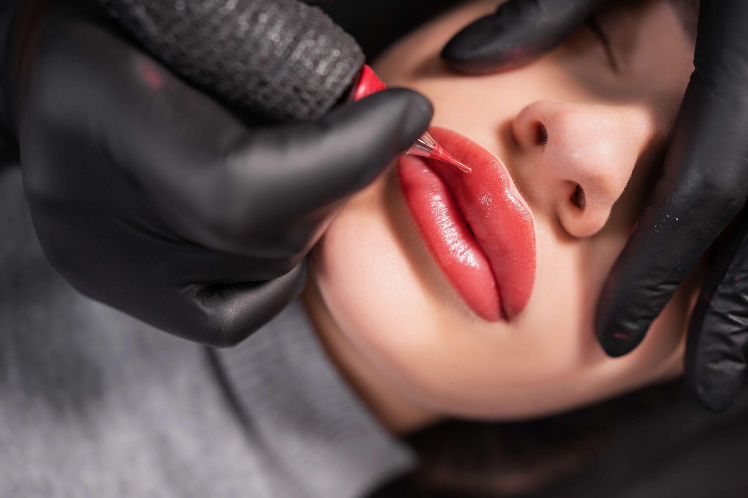 Rocío Martín - Experta en Micropigmentación en Madrid 9 Rocío Martín Micropigmentación Estética - Vista superior de una parte del rostro, donde unas manos enguantadas de negro utilizan una máquina de micropigmentación para aplicar pigmento en unos labios. Los labios ya tienen un color rosado rojizo intenso, indicando un proceso avanzado de micropigmentación de labios. La piel alrededor de la boca se ve limpia y clara.