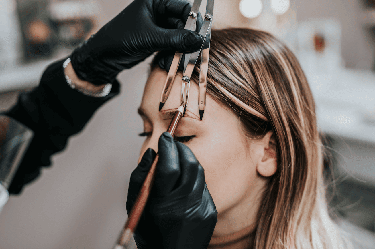 Diseño y visagismo Rocío Martín Micropigmentación Estética - Detalle de un primer plano de unas manos enguantadas en negro usando un calibrador de cejas y un pincel para delinear y medir las cejas de una mujer con cabello castaño claro. El calibrador de cejas se utiliza para asegurar la simetría y la forma perfecta de las cejas, mientras se aplica una marca o guía con el pincel