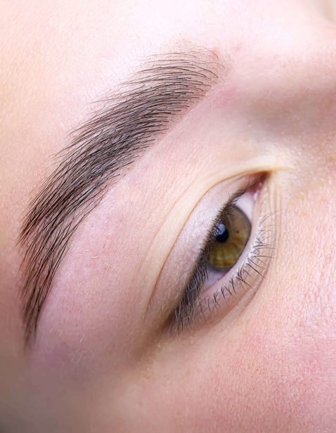 Rocío Martín Micropigmentación Estética - Primer plano de una ceja laminada, mostrando pelos individuales bien definidos y peinados hacia arriba, con un acabado brillante y pulido. El ojo adyacente tiene un iris de color miel, y la piel alrededor de la ceja está limpia y sin imperfecciones, destacando el resultado final del tratamiento de laminado de cejas.