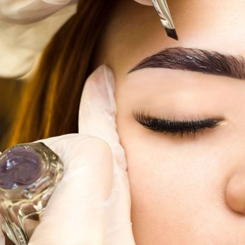 Rocío Martín Micropigmentación Estética - Detalle de un primer plano de unas manos enguantadas en blanco aplicando un producto oscuro con un pincel sobre la ceja de una mujer. Se observa el ojo cerrado de la mujer y pestañas largas y oscuras. A la izquierda, un pequeño recipiente de cristal con un producto de color púrpura oscuro, probablemente henna