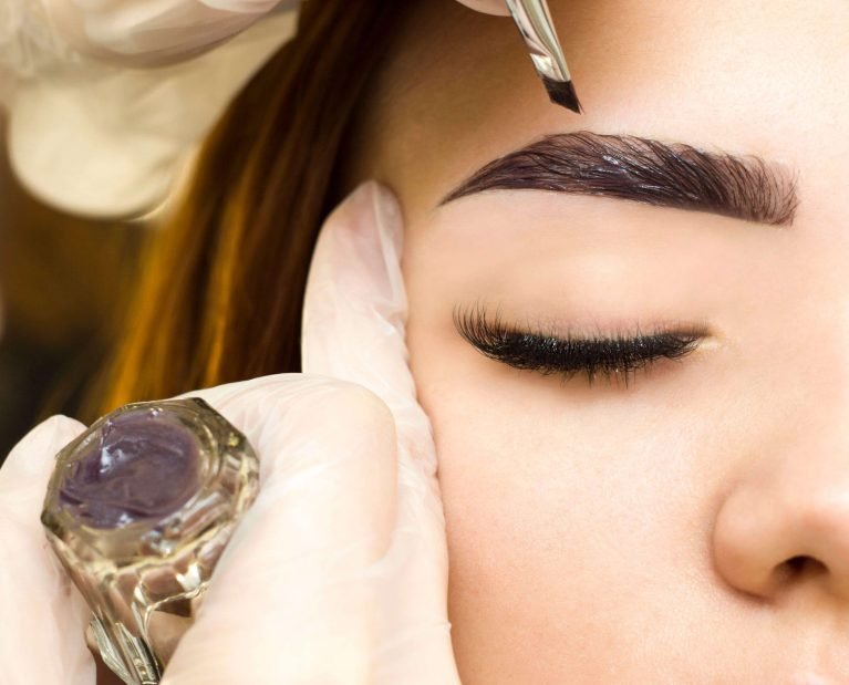 Rocío Martín Micropigmentación Estética - Detalle de un primer plano de unas manos enguantadas en blanco aplicando un producto oscuro con un pincel sobre la ceja de una mujer. Se observa el ojo cerrado de la mujer y pestañas largas y oscuras. A la izquierda, un pequeño recipiente de cristal con un producto de color púrpura oscuro, probablemente henna