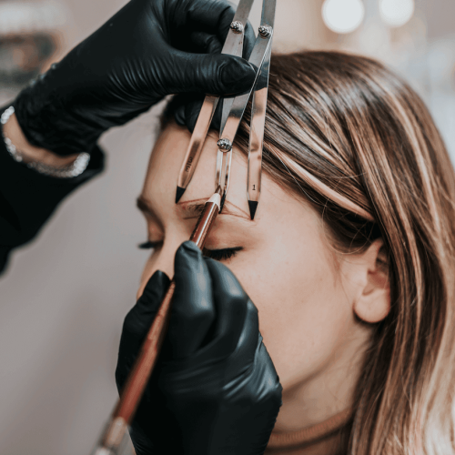 Rocío Martín Micropigmentación Estética - Detalle de un primer plano de unas manos enguantadas en negro usando un calibrador de cejas y un pincel para delinear y medir las cejas de una mujer con cabello castaño claro. El calibrador de cejas se utiliza para asegurar la simetría y la forma perfecta de las cejas, mientras se aplica una marca o guía con el pincel