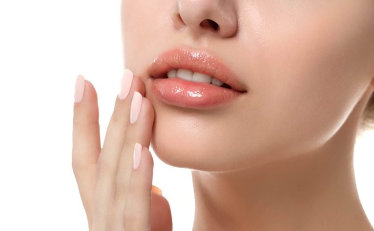 Rocío Martín Micropigmentación Estética - Primer plano de la parte inferior del rostro de una mujer, destacando unos labios voluminosos y brillantes con un tono nude rosado. Una mano con uñas bien cuidadas y esmaltadas en color rosa claro toca suavemente la barbilla. La piel de la modelo es clara y lisa